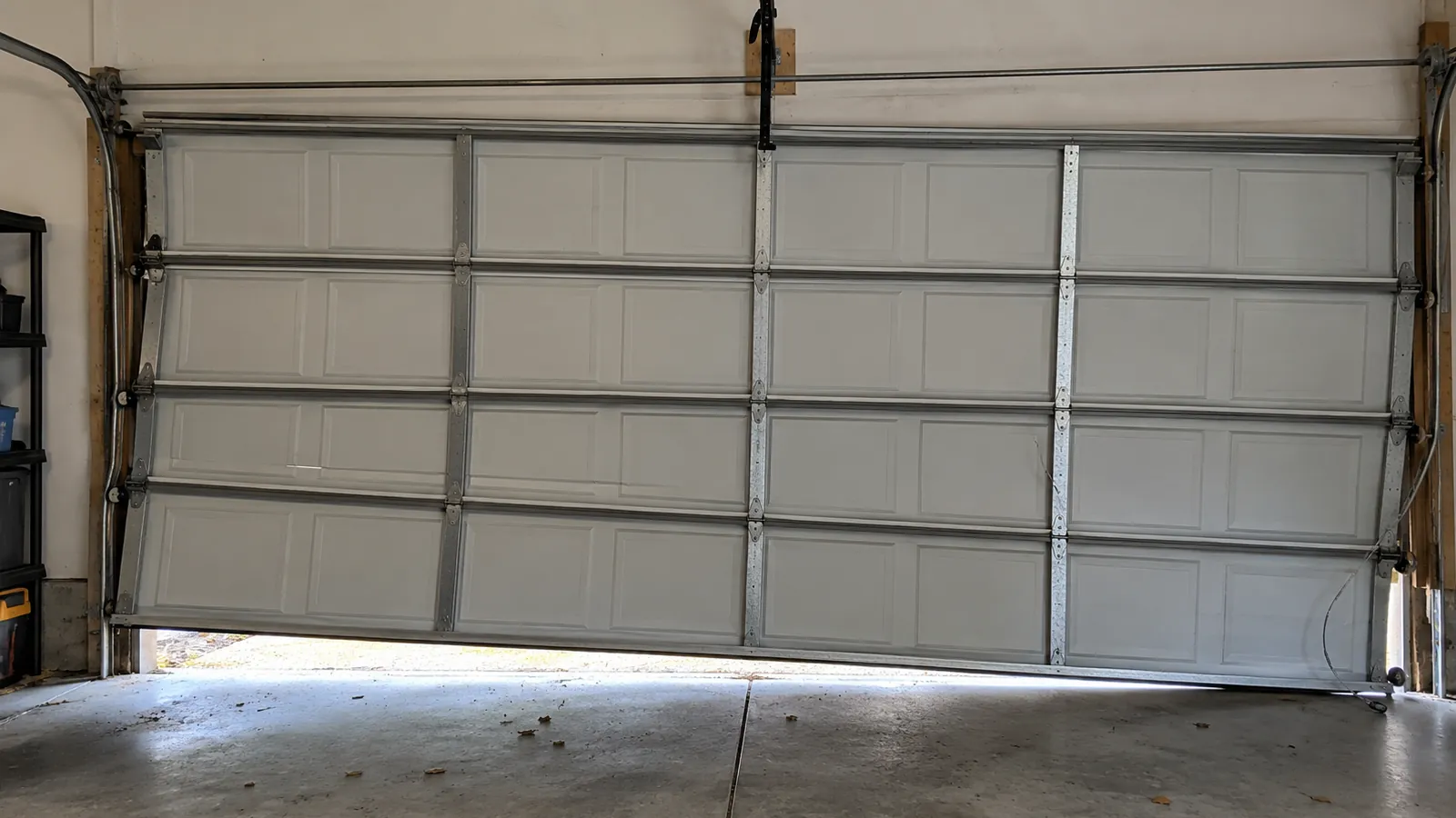 Two car garage door hanging unevenly in its opening, one side lower than the other, with a loose lift cable dangling near the bottom corner